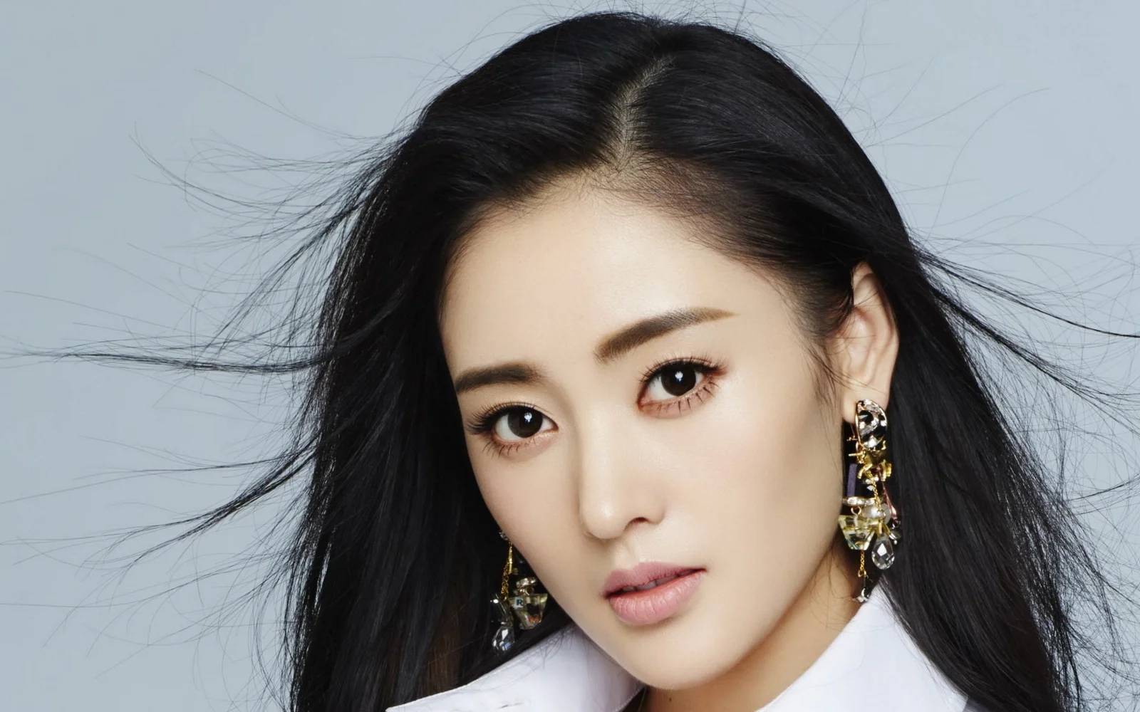 검은 머리의 여성이 장식적인 귀걸이를 착용하고 연한 회색 배경에서 카메라를 바라보고 있습니다.