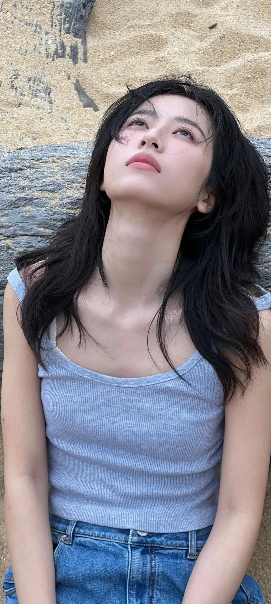 一名留着长黑发的女性穿着浅蓝色背心，靠坐在岩壁前抬头望向上方。