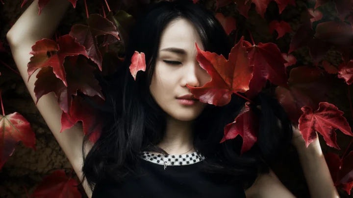 一名黑发女子被深红色叶片环绕，闭眼呈现宁静的肖像画面。