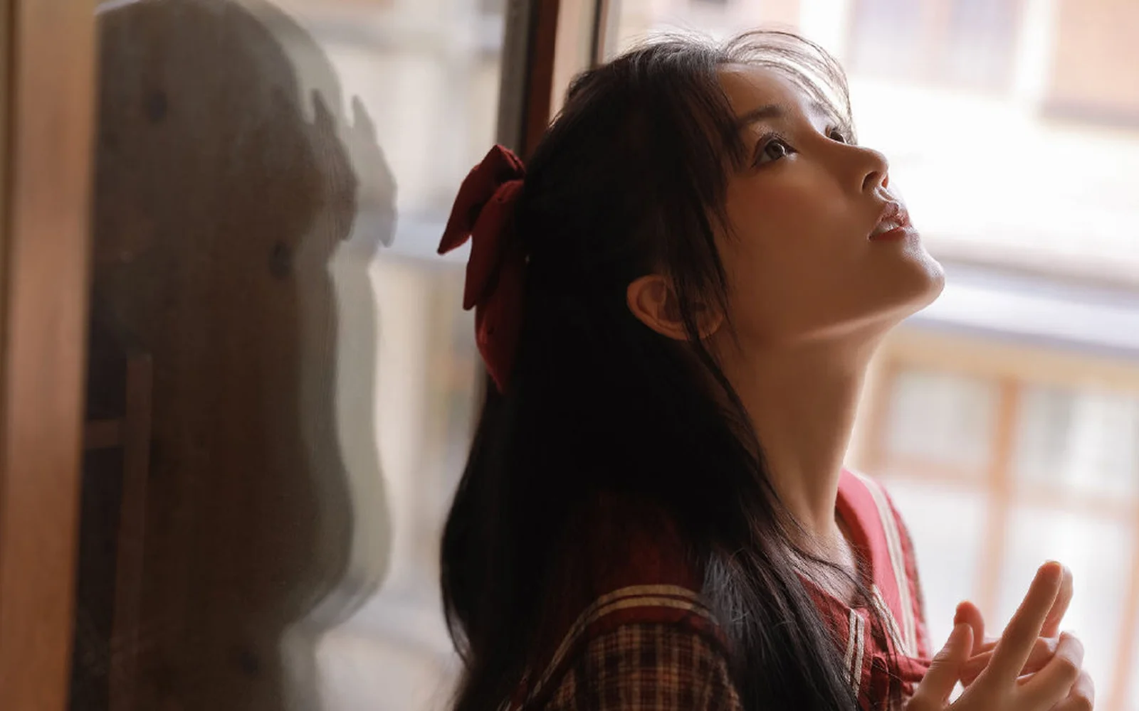 一名长发女子在窗边侧身抬头，柔和的窗光照亮了她的脸庞。
