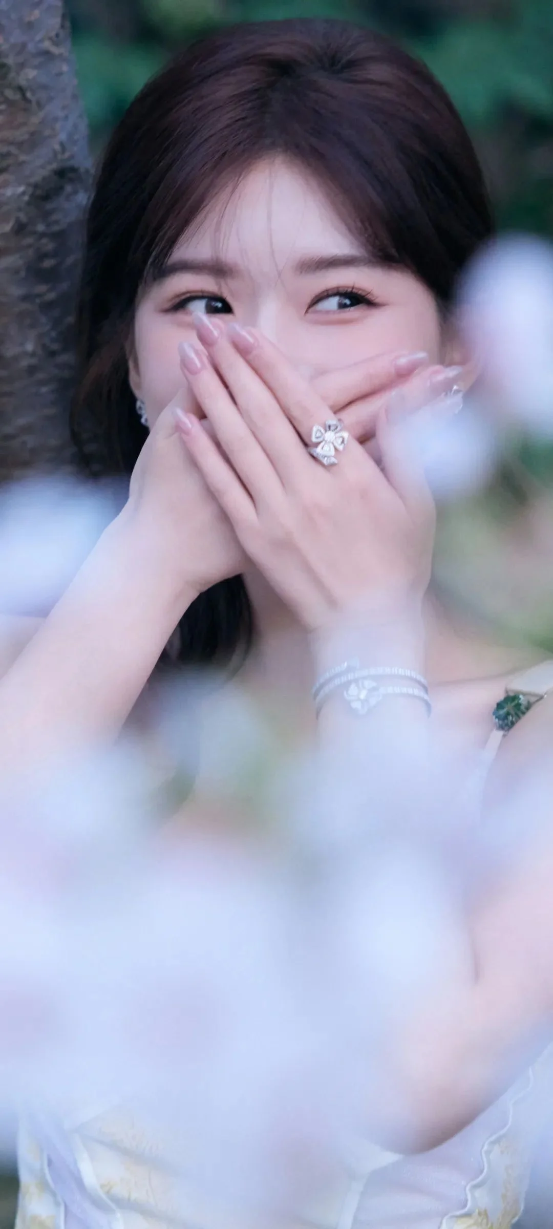 成人女性が木々と柔らかくぼけた背景の前で両手で口元を隠して微笑んでいる。