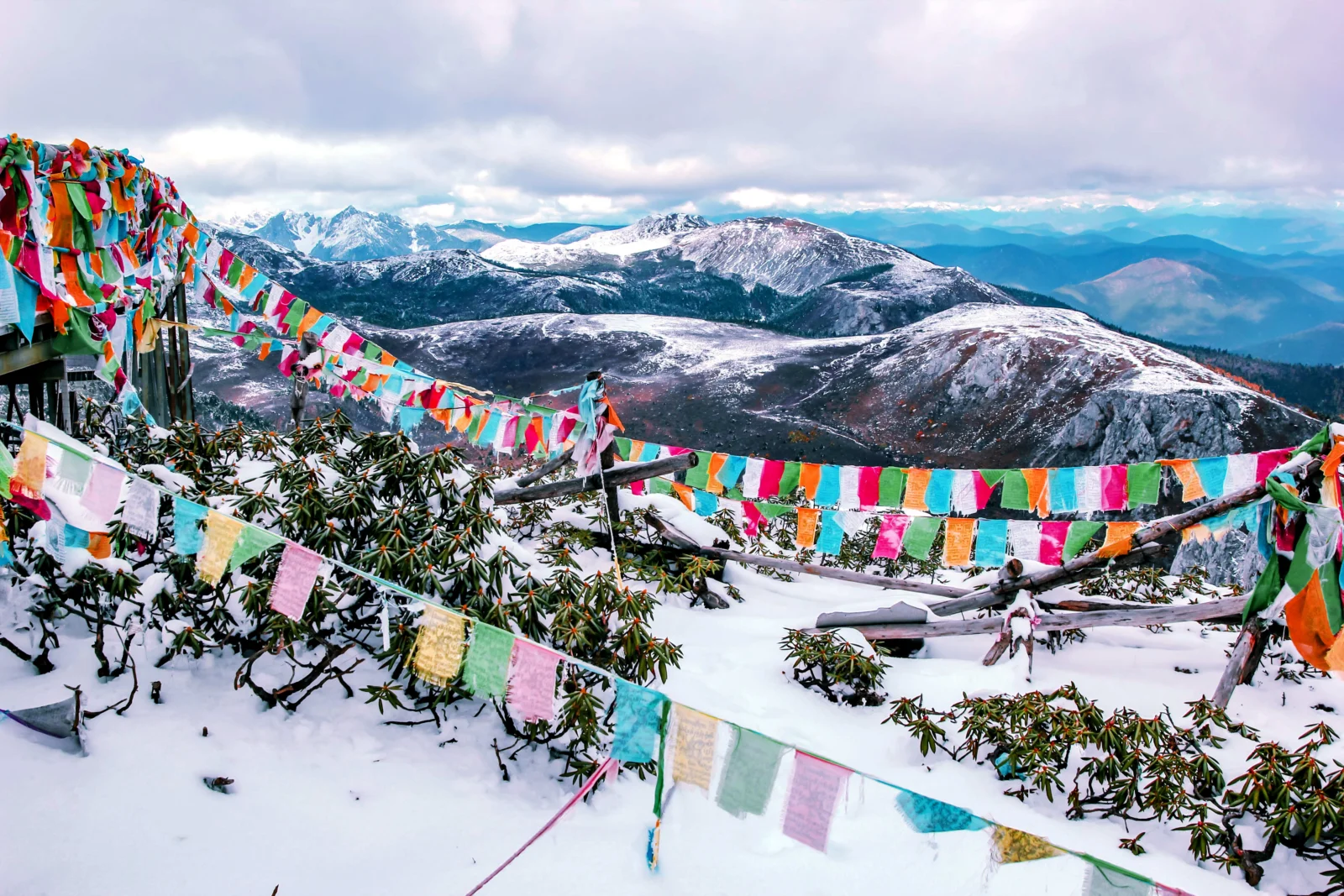 彩色经幡悬挂在积雪山坡前，远处是连绵的雪山和阴云天空。