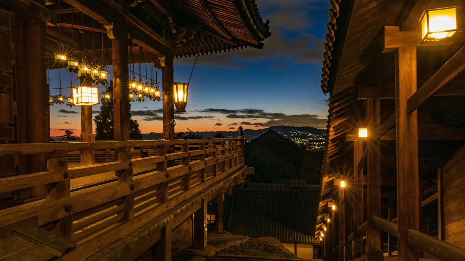 暮色中，木質建築與欄杆間懸掛的燈籠映照出溫暖光線，遠處山坡上散布著燈火。