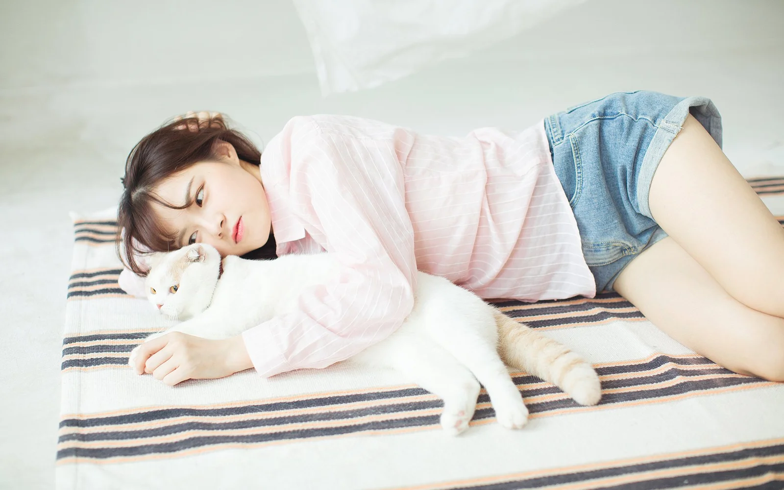 一名穿粉色上衣和牛仔短裤的女子躺在条纹床单上，身旁是一只白猫。