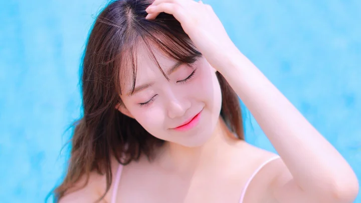 一名女性闭眼微笑，手扶头发，置身于明亮的蓝色背景前。
