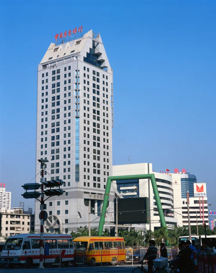 蓝天下，一座高层建筑矗立在繁忙街道旁，前景可见公交车和行人。
