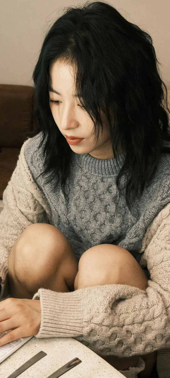 一名成年女子穿着针织毛衣坐在室内，低头看向一侧。