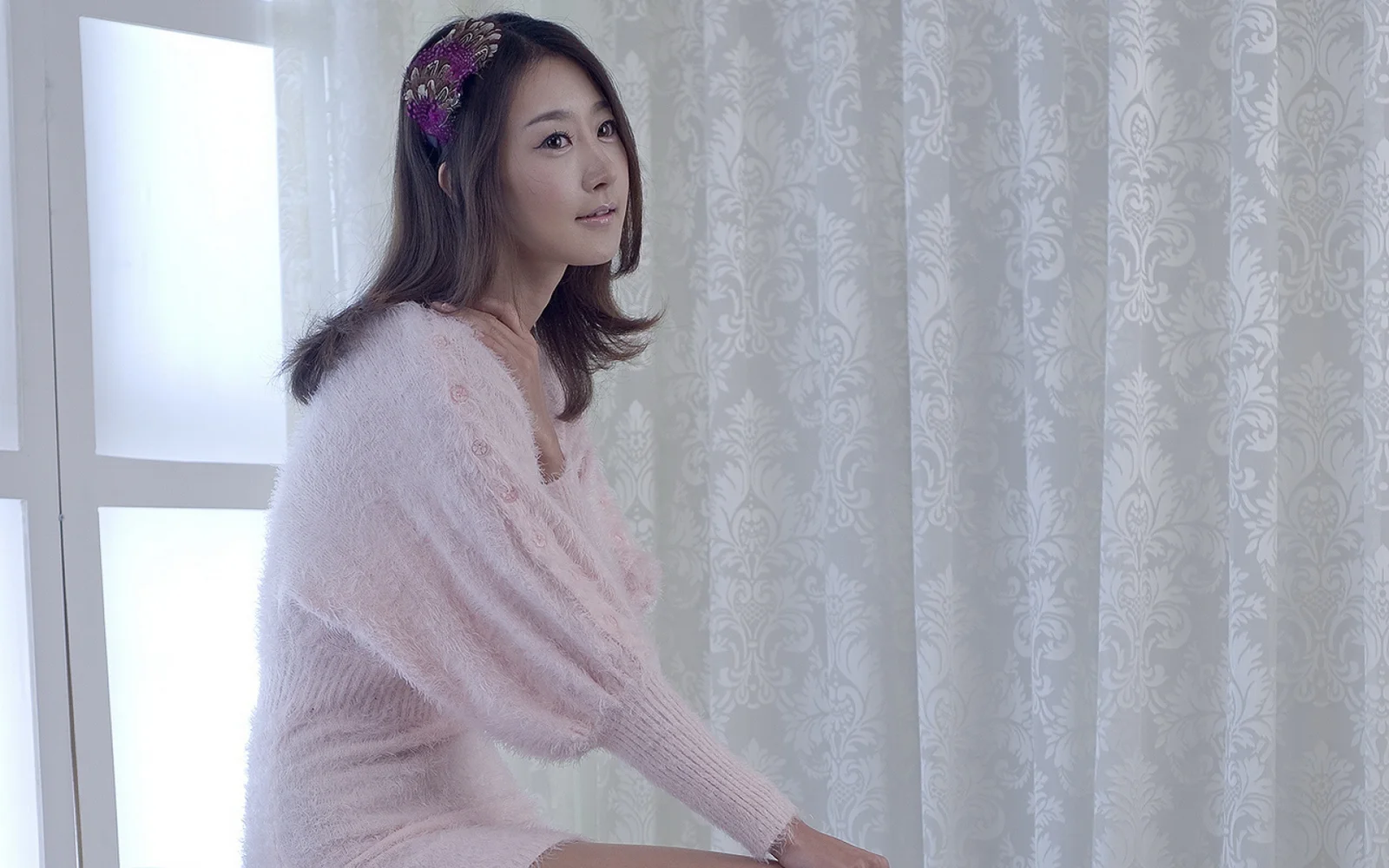 분홍색 스웨터를 입은 성인 여성이 밝은 무늬 커튼을 배경으로 창가에서 포즈를 취하고 있습니다.