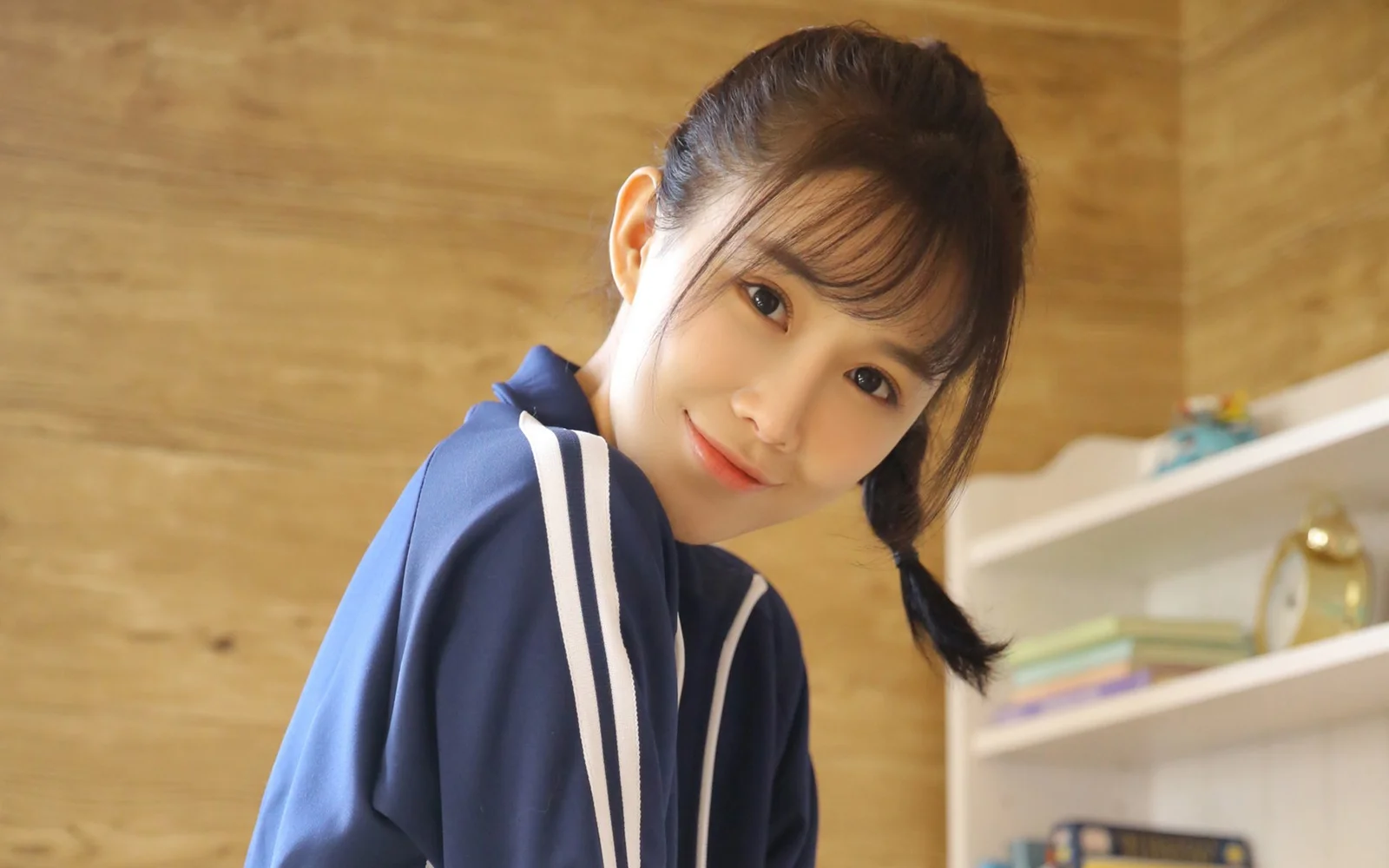 땋은 머리의 여성이 짙은 파란색 세일러풍 상의를 입고 나무 소재의 실내 배경 앞에서 카메라를 향해 미소 짓고 있습니다.