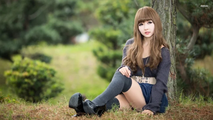 성인 여성이 나무 옆 잔디에 앉아 있으며 배경에는 부드러운 녹색 식물이 보입니다.