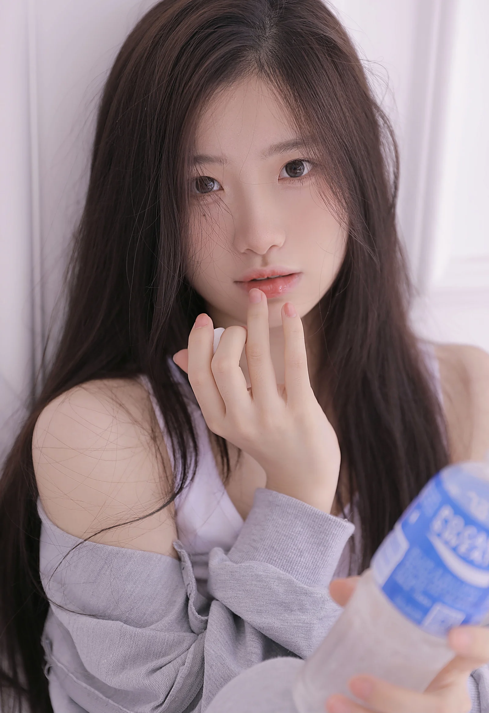 긴 머리의 여성이 입술에 손가락을 대고 파란색 병을 든 채 부드러운 실내 조명을 배경으로 카메라를 바라보고 있다.