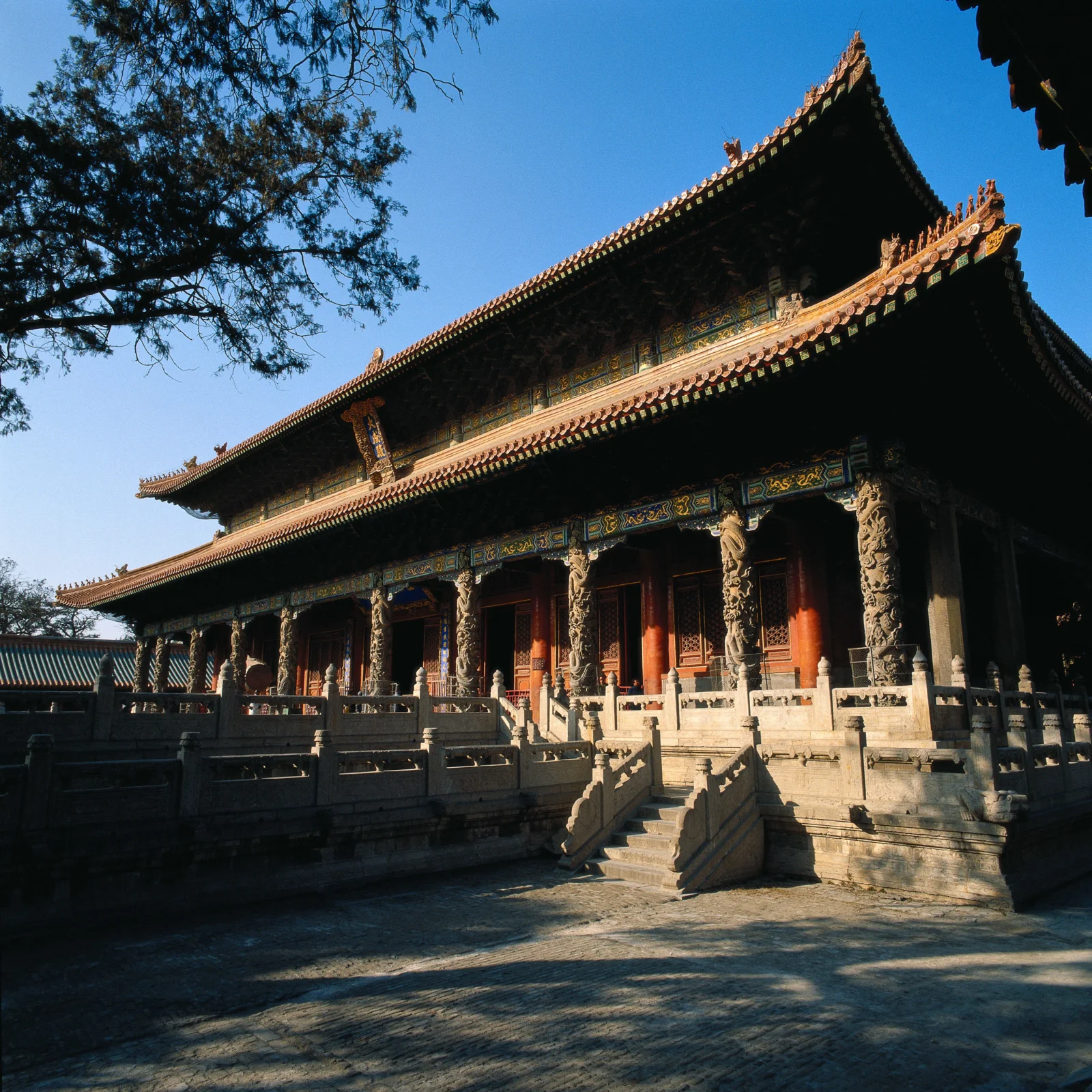 蓝天下，一座带有雕刻石柱和彩色屋檐的中国古建筑坐落在庭院中。