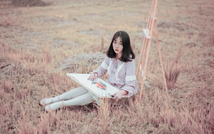一名女子坐在草地上，靠着画架描绘画板上的图案。