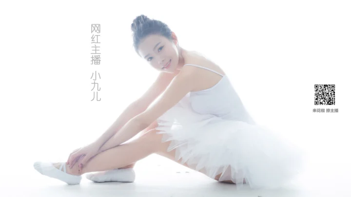 白いバレエ衣装を着た成人女性が、白い背景の前で座ってストレッチしている。
