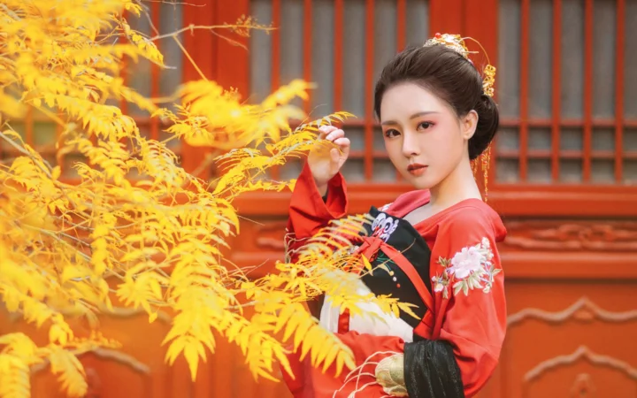 一名身穿红色传统服饰的女子站在金黄色秋叶前，背景是红色木质建筑。