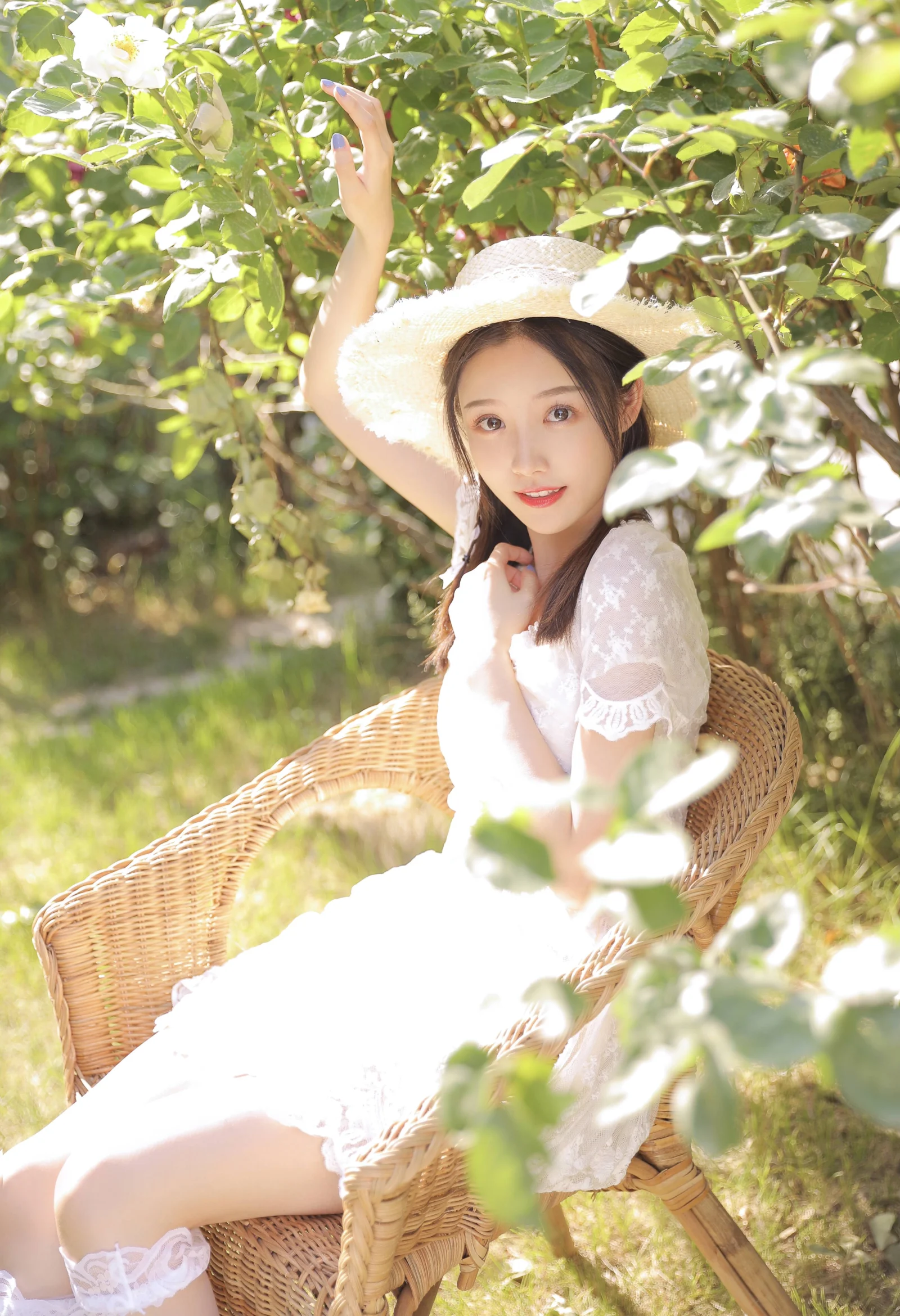 밀짚모자와 흰 드레스를 입은 여성이 햇살이 비치는 푸른 잎사귀 사이의 라탄 의자에 앉아 있습니다.