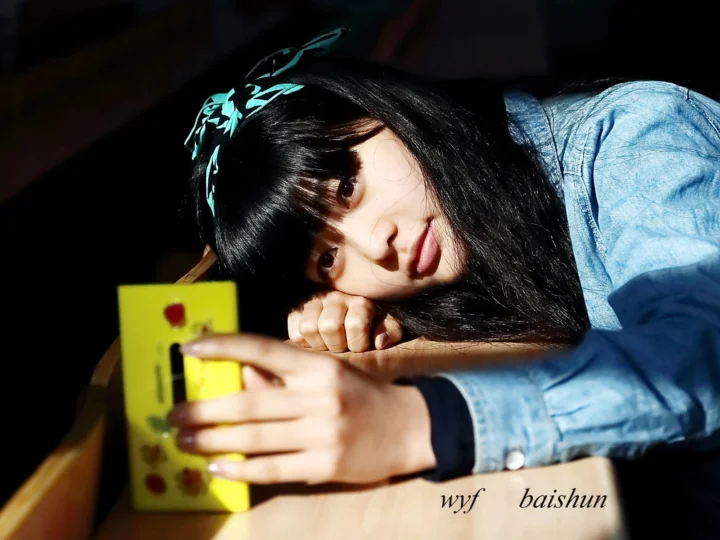 黒髪の女性が青いデニムジャケットを着て、テーブルのそばで横になりながら黄色いスマートフォンを見ている。