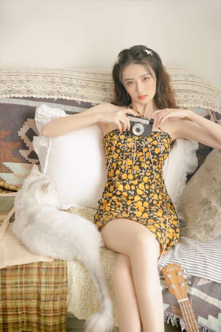 一位成年女性身穿黑底黄色花卉连衣裙，手持相机坐在铺有毯子和靠垫的沙发上。