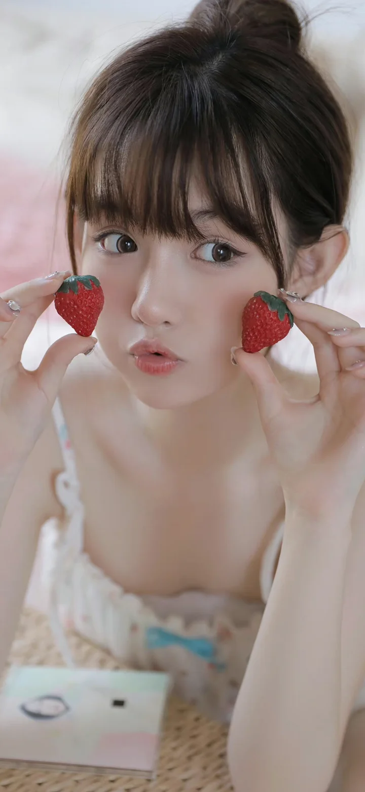 一名年轻女子手持两颗草莓靠近脸颊，面对镜头微微嘟嘴。