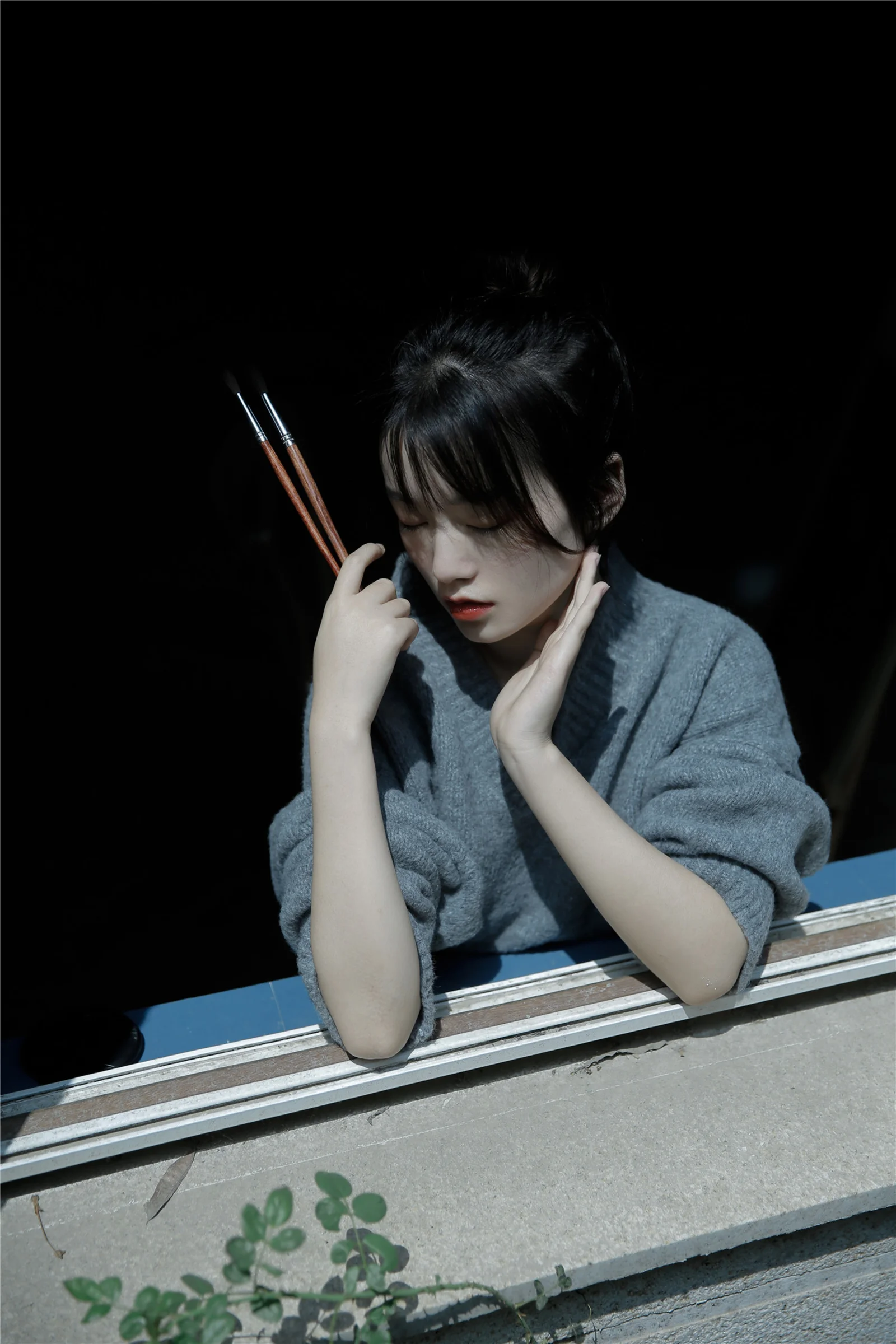 회색 스웨터를 입은 여성이 가느다란 젓가락 두 개를 들고 창가에 기대어 있으며 햇빛이 그녀와 콘크리트 창턱을 비추고 있습니다.