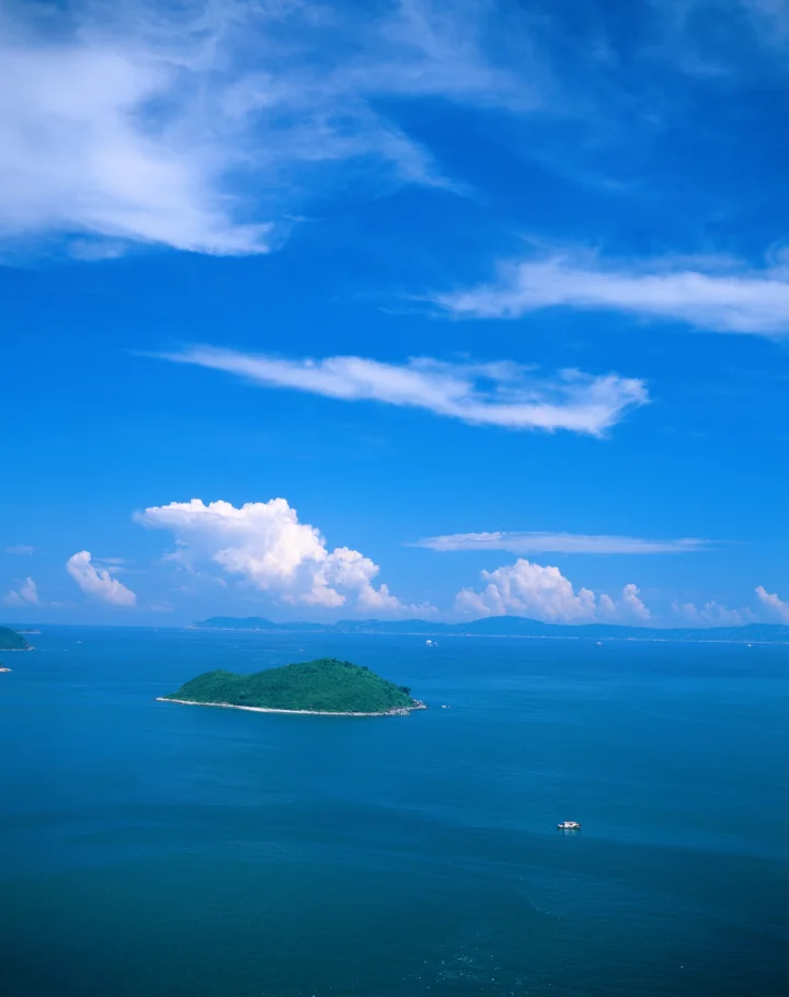 湛蓝海面上坐落着一座绿色小岛，远处的云层与晴朗天空构成开阔的海景。