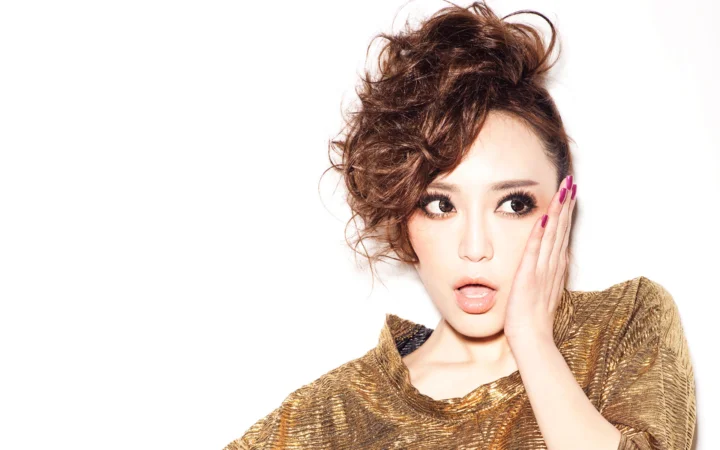 一名妆容精致、卷发造型的成年女性身穿金色上衣，在白色背景前摆姿势。