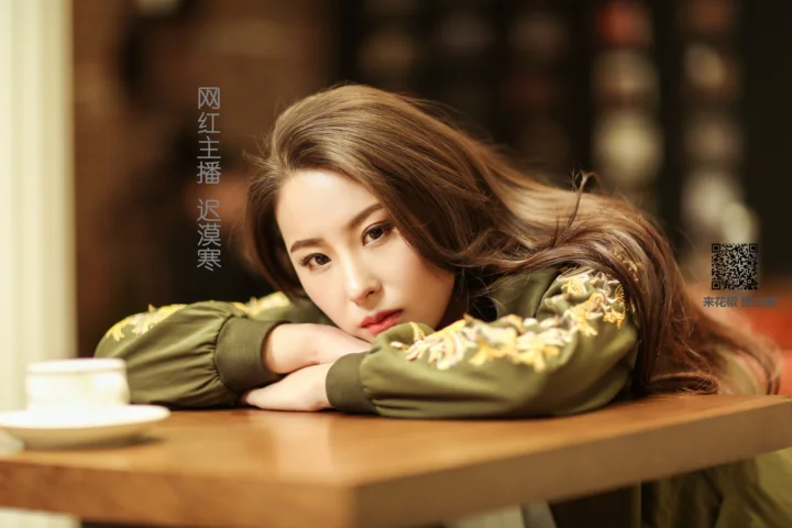긴 머리의 여성이 나무 테이블에 팔을 올리고 카메라를 바라보고 있다.