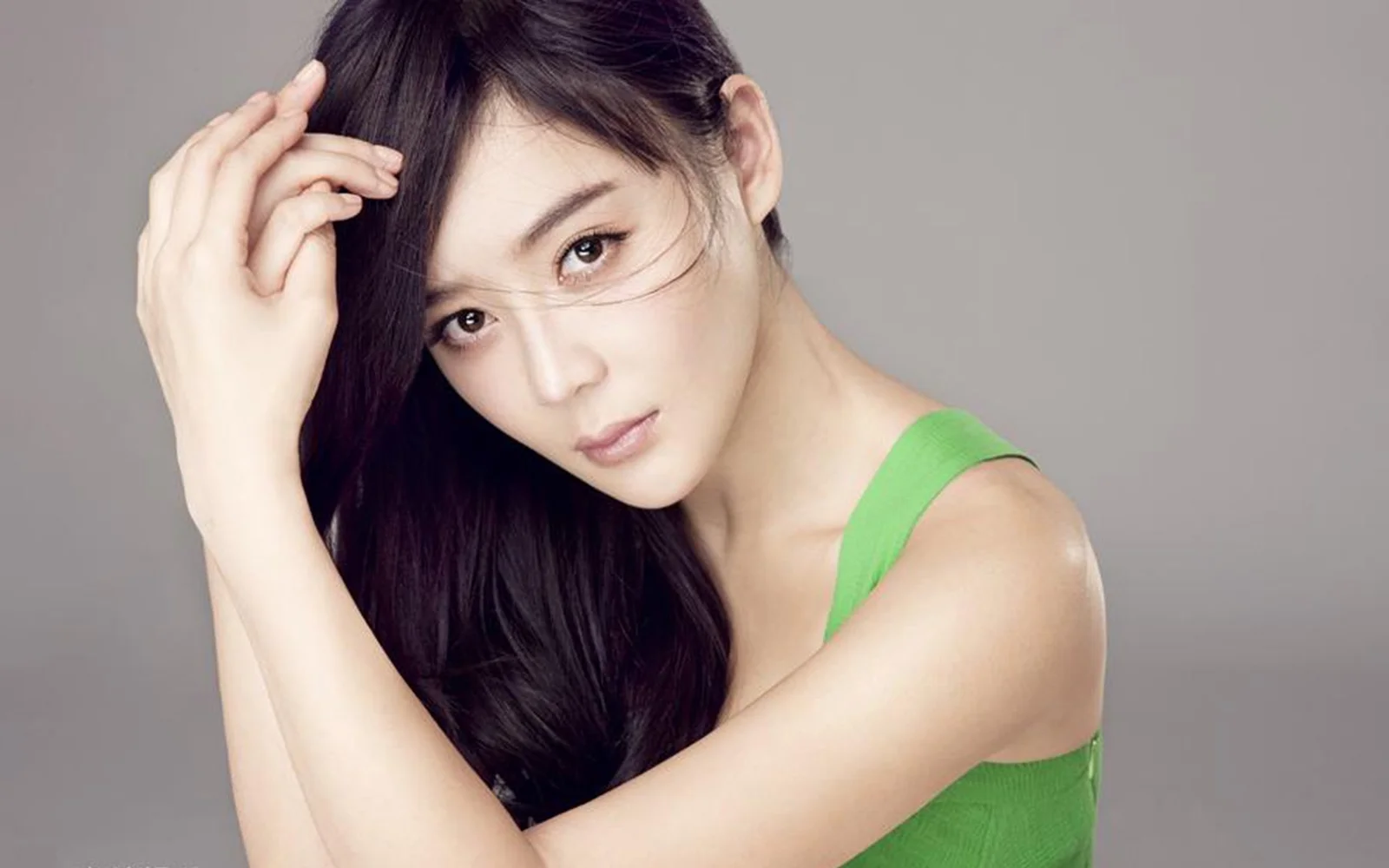 긴 머리의 여성이 초록색 민소매 의상을 입고 회색 배경 앞에서 포즈를 취하고 있습니다.