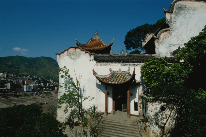 一座白墙灰瓦、带有翘角屋檐的传统中式建筑坐落在山城景色前。