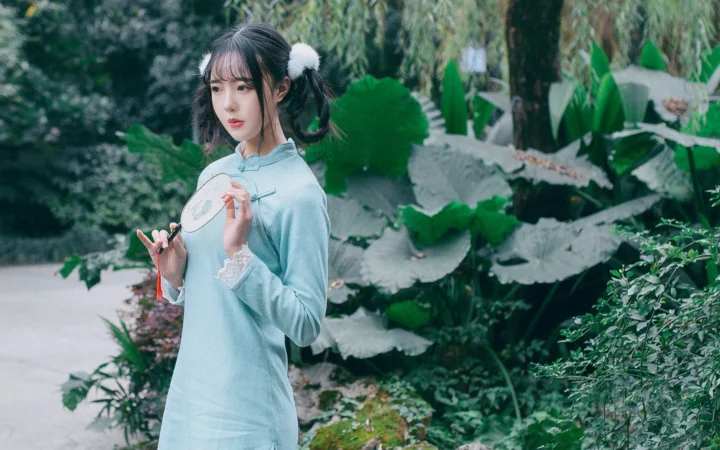 一名身穿浅蓝色传统服饰的女子站在绿意盎然的庭院中，手持团扇。