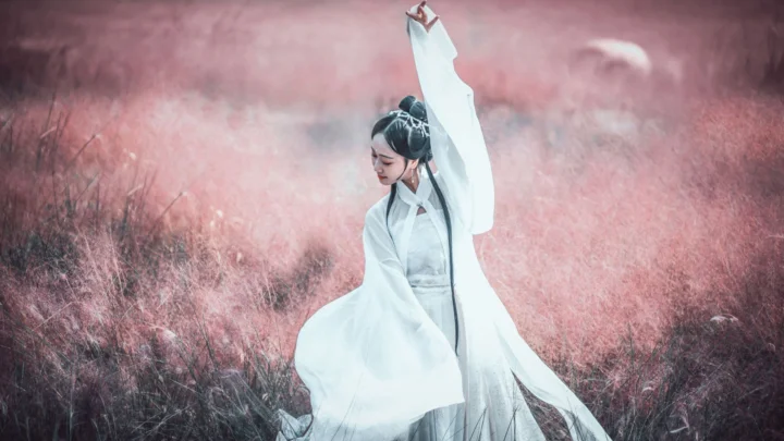 一名身穿白色长袍的女子站在粉色草地中，抬起一只手臂。