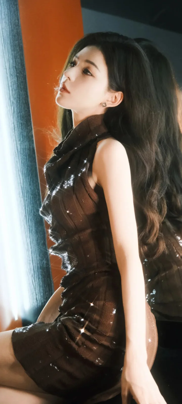 一名成年女性侧身坐在室内窗边，穿着黑色亮面无袖连衣裙，长发披落在肩后。