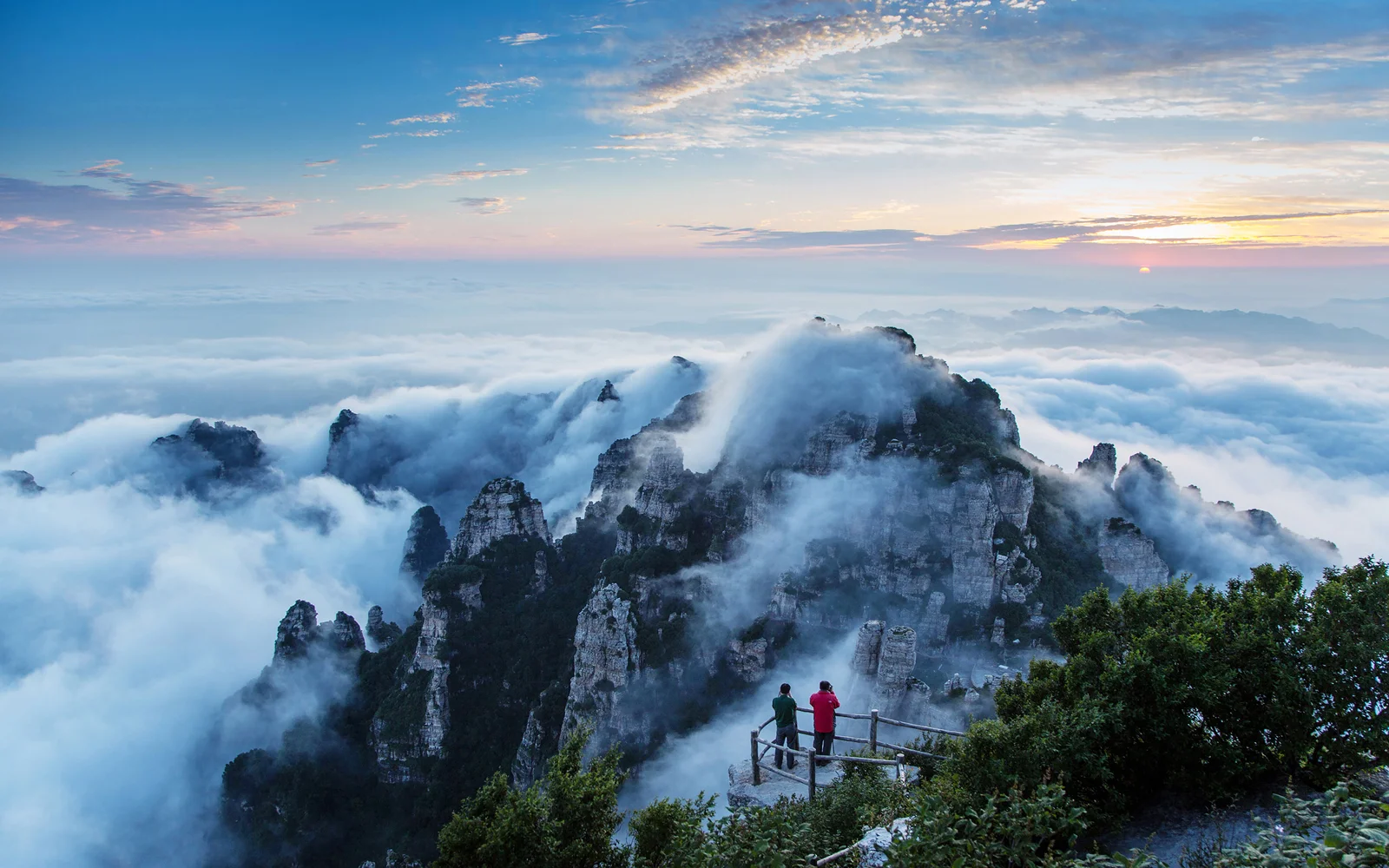 云雾缭绕的群山延伸在日出天空下，前景观景台上可见两名远处行人。
