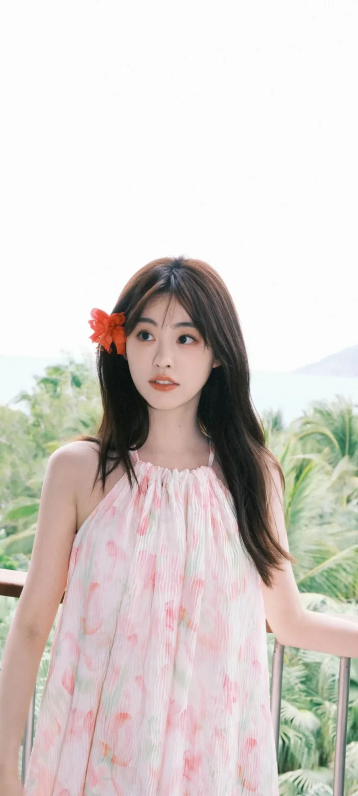 一名女子穿着粉色连衣裙站在户外栏杆旁，头发上别着一朵红花。