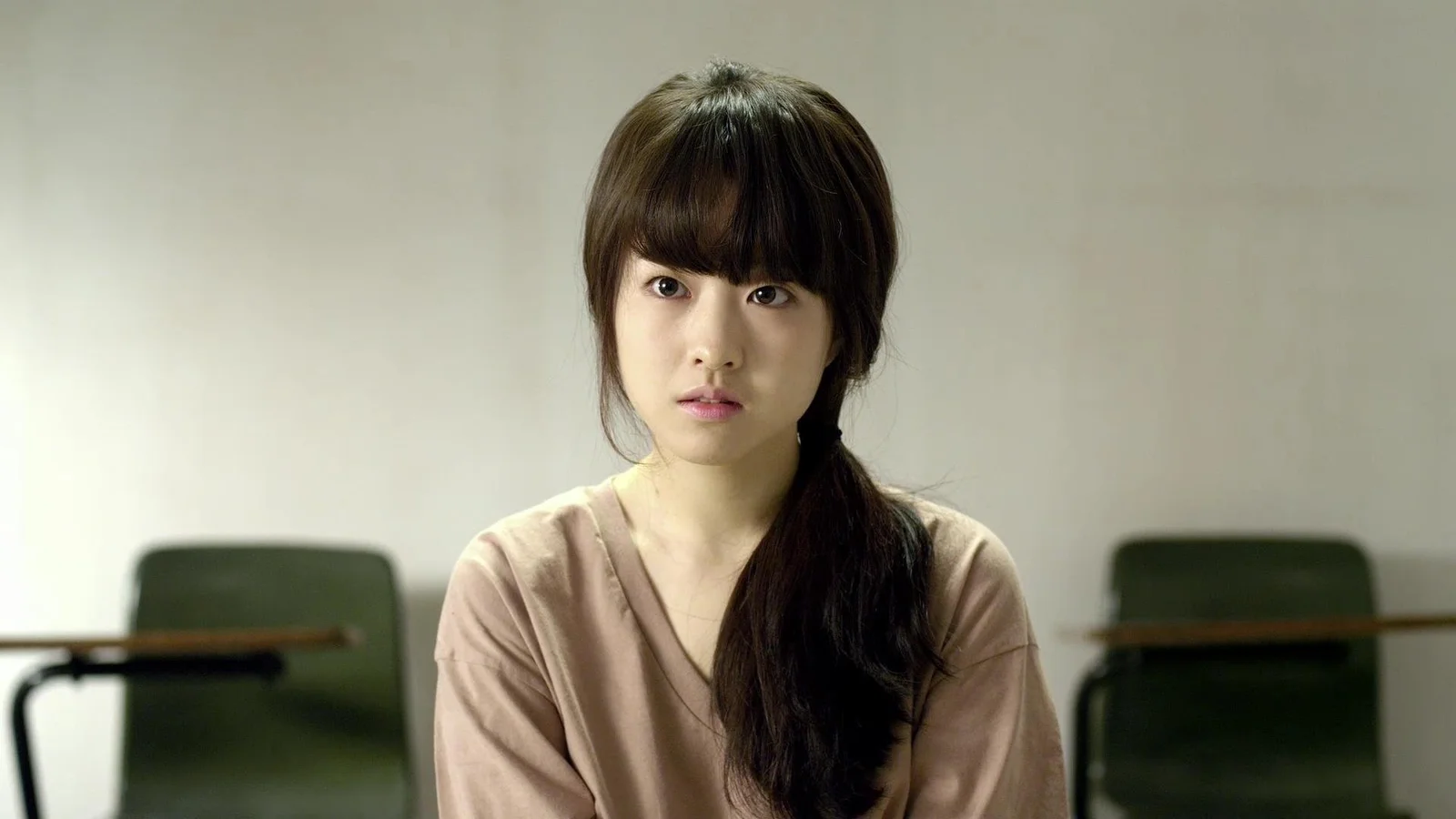 긴 머리의 여성이 실내에 앉아 있고 뒤로 의자와 테이블이 보입니다.