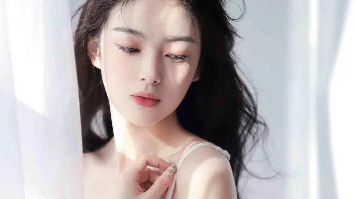 一名成年女性站在明亮窗边，低头触碰肩带，柔和光线照亮她的面容。