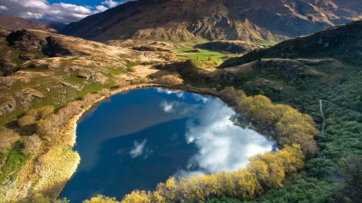 群山环绕的山谷湖泊映照着天空中的云朵，周围是金色植被与岩石地貌。