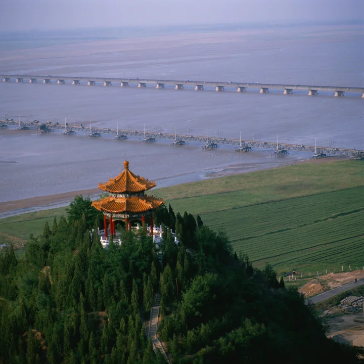 一座传统亭阁坐落在绿树间，远处是宽阔河流、长桥和整齐的农田。