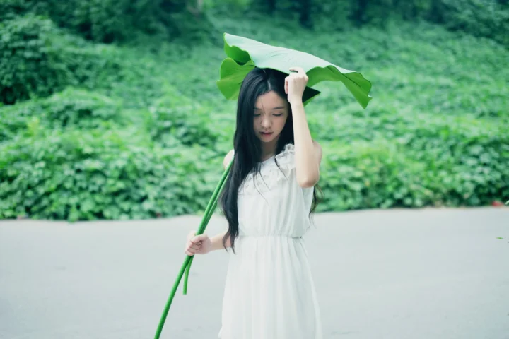 一名身穿白裙的女子在绿色户外环境中举着荷叶遮在头顶。