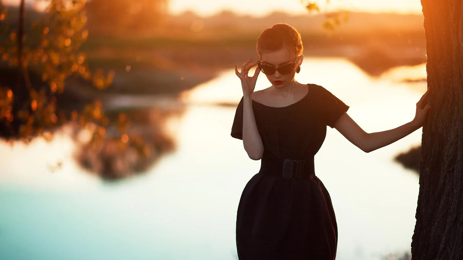 サングラスと黒いワンピースを身に着けた女性が、夕日に照らされた湖畔の木のそばに立っている。