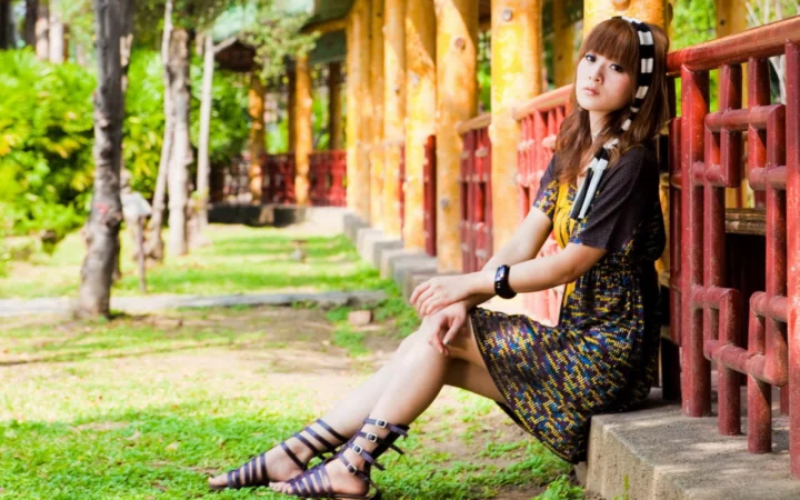 一名女性坐在户外木制栏杆旁，身后是树木、草地和木柱。