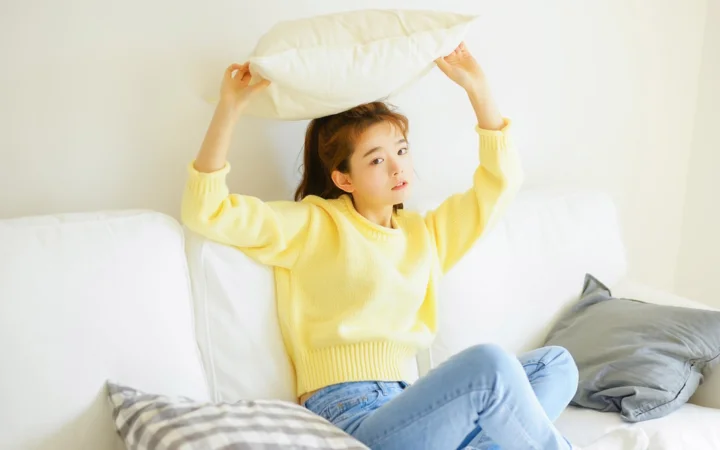 一名穿黄色毛衣的女性坐在白色沙发上，举着白色枕头。
