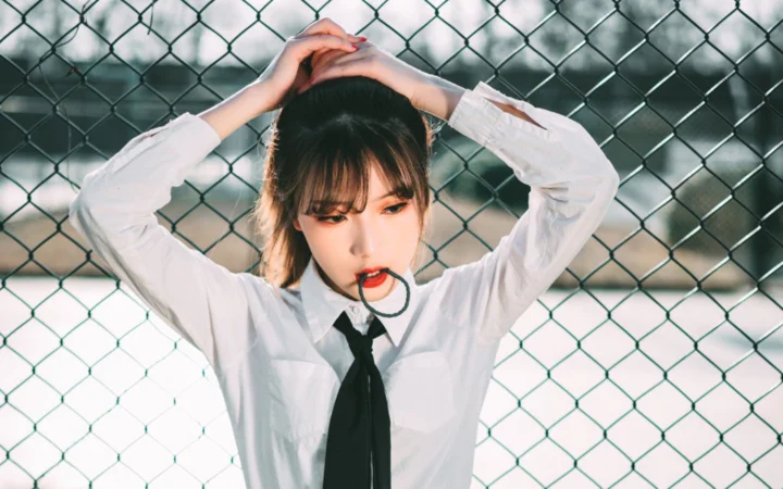一名穿白衬衫和黑色领带的女性站在链式围栏前，双手举过头顶并咬着一根黑色环状物。