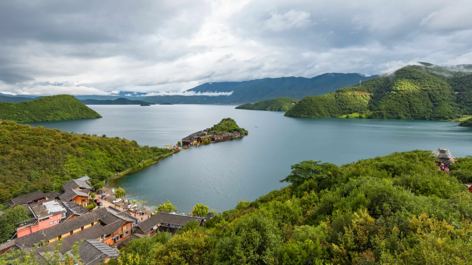 群山环绕的湖泊延伸在绿色山林之间，前景可见湖畔建筑，天空覆盖着厚重云层。