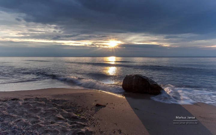 夕阳穿过云层照在平静的海面上，海浪轻拍着沙滩和岸边的岩石。