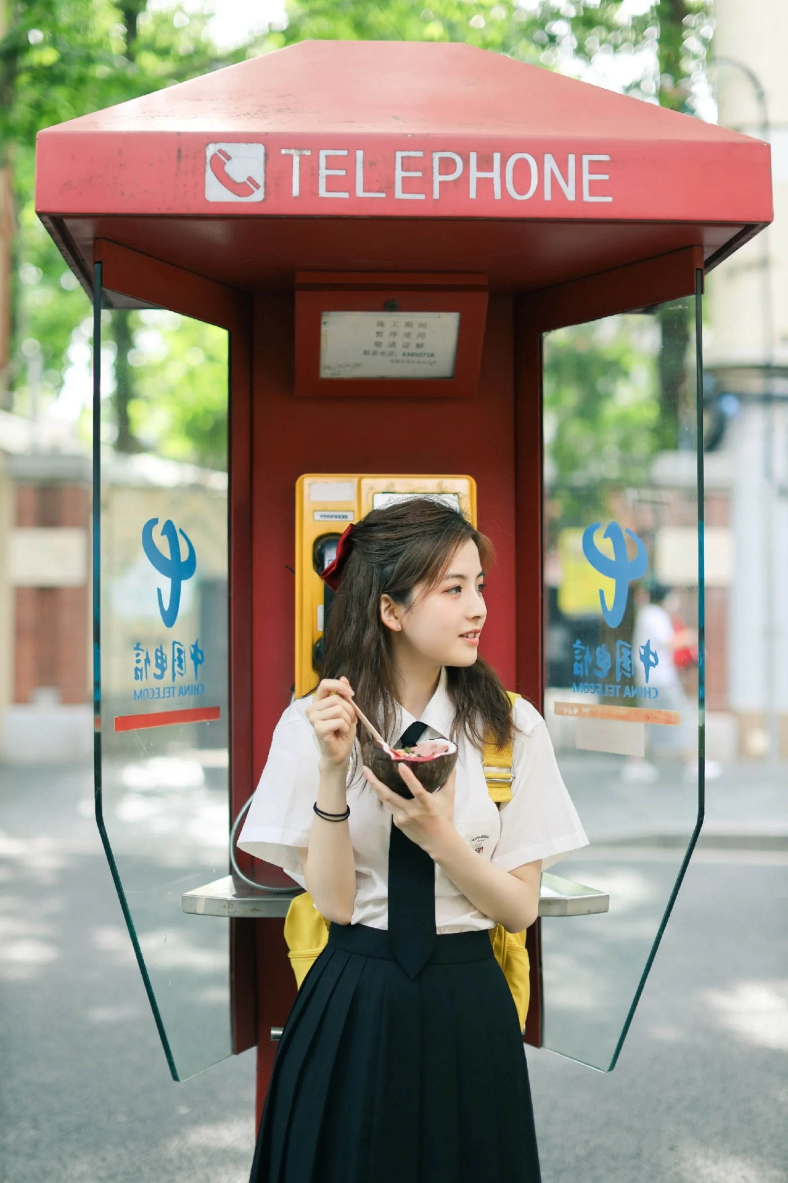 한 여성이 야외의 빨간 전화 부스 안에서 숟가락과 그릇을 들고 서 있다.