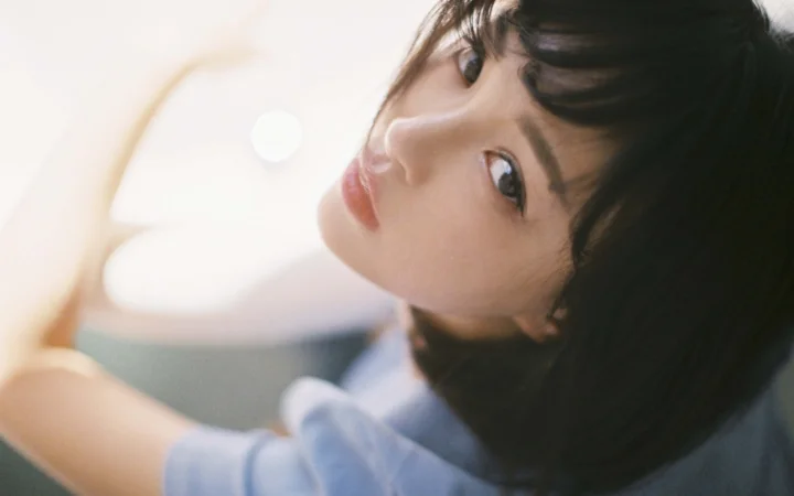 검은 머리의 여성이 부드러운 빛 속에서 고개를 기울여 카메라를 바라보고 있다.
