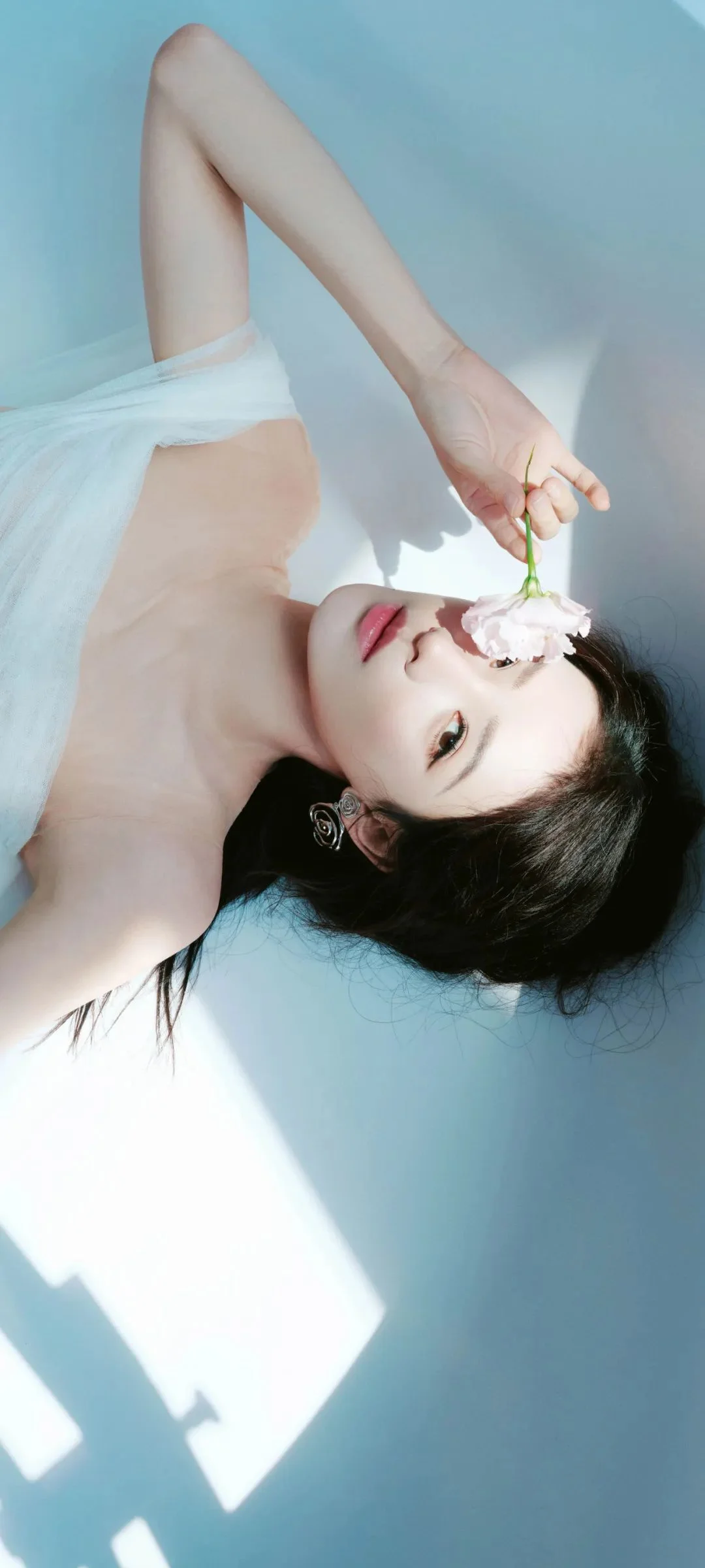 一名成年女性身穿白色服装躺在浅蓝色背景上，手持一朵白花靠近脸庞，画面带有明亮的光影。