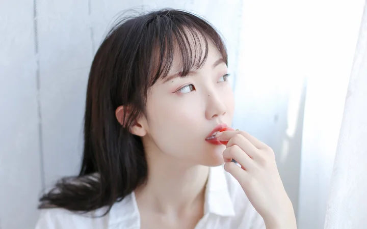一名女性站在窗边，手持唇彩靠近嘴唇，柔和的自然光照亮她的侧脸。