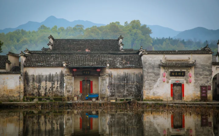 灰瓦白墙的传统村落建筑倒映在池塘中，远处可见群山与树木。
